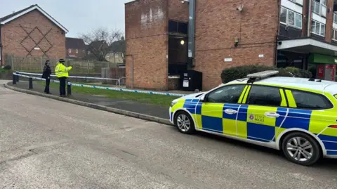 An Essex Police branded car parked on a pavement by a road. There is a cordon around a house and two police officers standing in front of it 