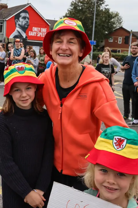 Getty Images Laura McAllister wearing a bucket hat standing with two children also wearing  bucket hats. There are crowds behind her cheering as runners go past from the Cardiff Half Marathon with a red mural of Gary Speed seen on a wall to the right side of the image.