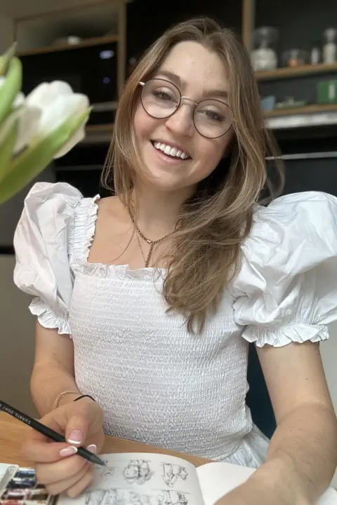 Lucy Dunbar A woman with long mousy brown hair falling down one shoulder is wearing circular, thin-rimmed glasses and smiling into the camera. She is wearing a dainty gold necklace with a chain going down the middle and a white top with puffed shoulders. She has a pencil in her right hand and you can make out small intricate pencil drawings on paper below it, next to a watercolour pallet. Behind her is a vase of white flowers and shelving.