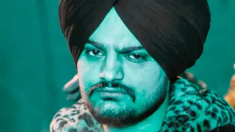 Sidhu Moose Wala stares into the camera in a promotional shot - he wears a black turban and has a leopard skin collar 