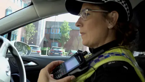 A uniformed police officer wearing glasses listening to a walkie talkie.