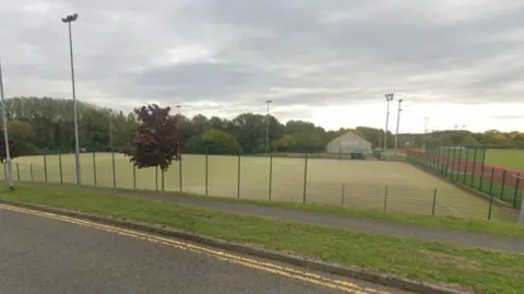 Google An image of an artificial football pitch surrounded by fencing.  It is bordered by trees to the left. There is a house in the background and an athletics track to the right. Floodlights on stands are visible in the background.