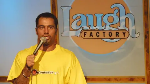 Getty Images Joe Rogan, wearing a yellow shirt with a black logo that reads 'dankster', speaks holding a microphone during a stand-up set at the Laugh Factory in Hollywood in 2001