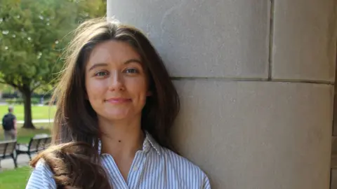 Clíodhna Ní Chorráin Clíodhna Ní Chorráin, who has long brown hair and is leaning against a wall. She is smiling and is wearing a striped blue and white shirt.