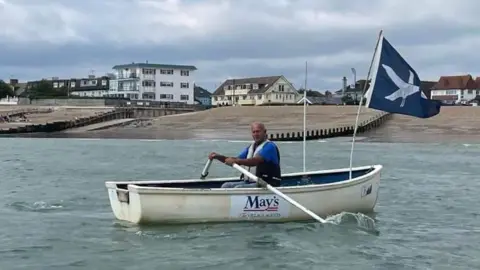 Ross Fisher A man wearing a blue shirt and white life jacket rowing a white dinghy boat. The boat is flying a blue flag with a white dove on it. The boat is in the sea with the beach and houses in the background