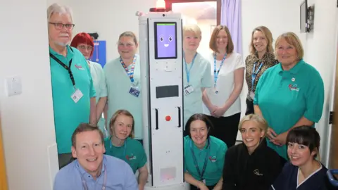 South Tees Hospitals NHS Foundation Trust James Dunbar with the robot and a group of about 11 other people stand around a robot cleaner which has a purple smiley face on its screen