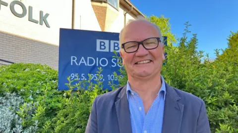 Vikki Irwin/BBC Matthew Hicks is standing in front of the BBC Radio Suffolk building. There is a hedge to the right in the background and to the left there is a BBC Radio Suffolk sign. Matthew is wearing a bright blue shirt and dark blue coat. He is smiling and looking directly at the camera. 