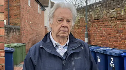 A photo of a man stood in a street with bins on either side of him. He has white hair and is wearing a blue coat and a blue and white top underneath