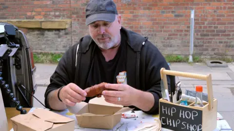 Stephen Peel A man wearing black clothes and a cap sits at a table outside holding a battered sausage. A cutlery holder next to him has the words 'Hip Hop Chip Shop' written on the front.