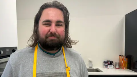 A young man with long brown hair and a full beard smiles with closed lips as he stands in a kitchen with white walls, a boiler and a sink. He is wearing a grey jumper and a bright yellow apron.