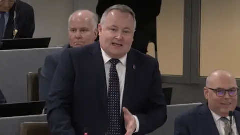 Senedd Cymru Darren Millar stood in the debating chamber wearing a dark suit, dark tie with white spots and gesturing in front of him. Paul Davies is sat next to him on the right and is wearing glasses.