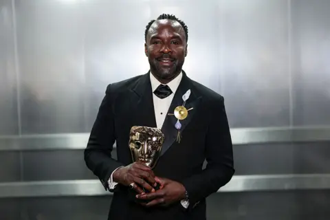 Getty Images Ariyon Bakare in a black suit holding his Bafta award