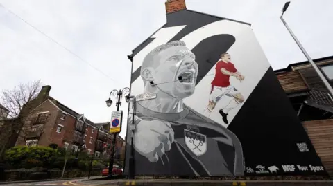 EPA A large black and white mural shows Paul Mullin punching the air and shouting during a game. A colour painting of Mullin playing can be seen to the right of the mural. It is on the gable end of a building. A multi-storey building can be seen across the road on the left.