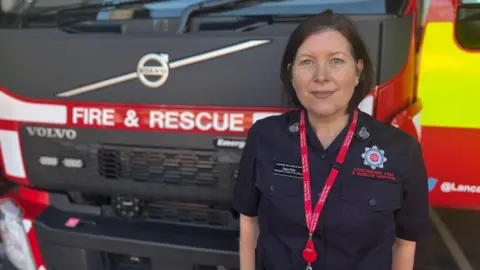 Samantha stops in front of a fire truck. She uses a Navy Blue Fire and Blue Rescue Uniform from Lancashire and a red cord. She has balanced brown hair and smiles at the camera.