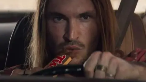 A man with long hair and a moustache drives a car in a close up picture. He eats a Twix bar at the same time.