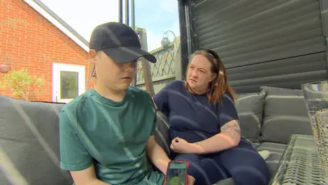 Tora sitting on an outdoor sofa in the garden with her son Mason. Mason is wearing a black baseball cap and green t-shirt and is holding a phone. Tora is wearing blue and is looking at Mason.