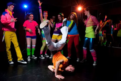 A group of young people dressed in 80s gear dancing with a young man breakdancing in the front