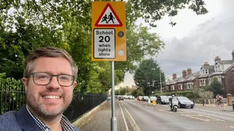 Rob Colwell A man wearing glasses smiles towards the camera next to a flashing speed limit sign outside a school, with cars and a motorbike passing by