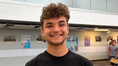 jack is smiling for the camera in a school passagew2ay , He has curly brown hair and stubble. 