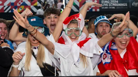 Una mujer en Pensilvania sostiene una bandera de Inglaterra alrededor de su cabeza y extiende los brazos hacia arriba mientras usa gafas verdes.