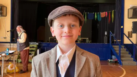 A school boy in costume. He has a flat cap on and a light coloured jacket. He is standing in front of a stage in a school hall
