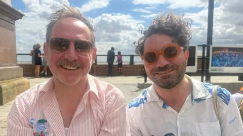 Two men in summer shirts are wearing sunglasses and smiling into the camera as they are sat down in along a marina. The man on the left is wearing a red and white stripy top and the man on the right is wearing quirky orange sunglasses with a white shirt with blue trees on it.