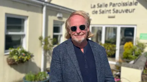 Bob Murray stood infront of St Saviour's Community Centre. He is wearing sunglasses, a blue top and a grey patterned blazer. He has long blonde hair and a blonde and grey beard.