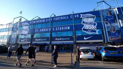 The front of a football stadium. The entrance to St Andrew's Park, the Birmingham City ground. The building is blue with large windows and a sign reading "Birmingham City Football Club". People are walking on the pavement near the entrance and cars are parked on the right.