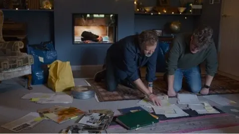 Hardy Pictures Two men in casual shirts, jumpers and jeans kneel on a grey carpeted floor looking through what is clearly an extensive archive of old papers and photos. 