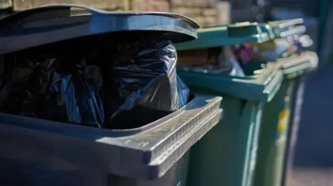 Getty Images A black recycling bin with its lid open to reveal a black binbag is in the foreground. In the background are two green recycling bins, with bags peeking out of the slightly opened lids.