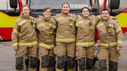 Five women in fire uniforms stand arm-in-arm