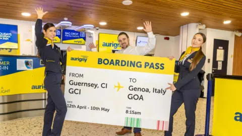 Three Aurigny employees hold a large boarding pass. The yellow, white and blue boarding pass states: "From Guernsey to Genoa Italy". The workers are wearing uniforms and are standing inside an airport.