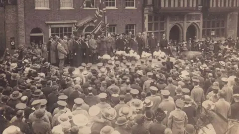 A black and white image of a two-minute silence in Farnham on 1 May 1916 with crowds of people and dignitaries with flags on stage in front of a big building
