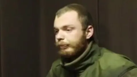 A man believed to be James Scott Rhys Anderson, 22, a former British Army soldier captured while fighting for Ukraine, is seen wearing green military clothing and sat in front of a plain brown background.