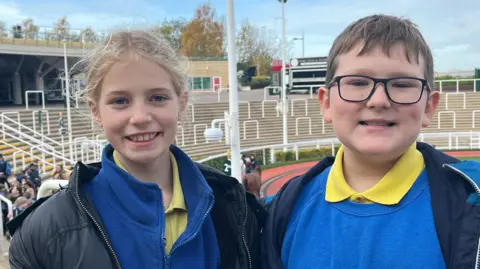 Two school children, one girl with blonde hair and one boy with short brown hair and glasses, are wearing royal blue jumpers and yellow polo tops. They are standing in front of the parade ring at Cheltenham Racecourse. They are both smiling. 