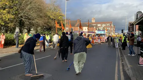 People wearing traditional clothes sweeping a tarmac road with crowds and a traditional float seen in the background