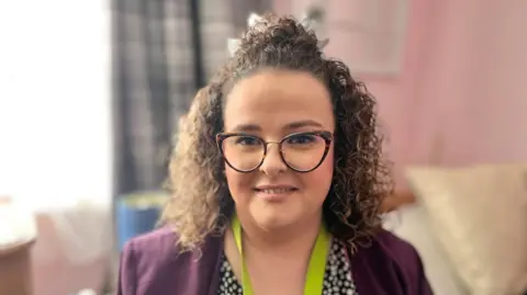 Martin Giles/BBC Rhiannon Reed is wearing a dark purple cardigan and blue and white spotted dress. She is smiling at the camera and is wearing glasses. She is sitting in a room with a bed in the background. 
