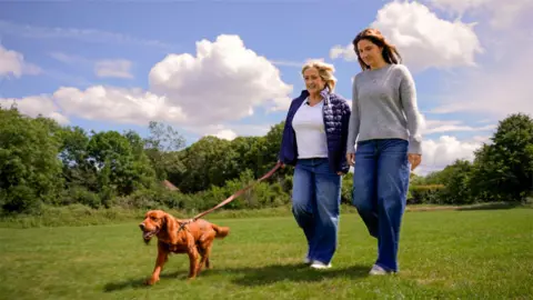 Two people walking across a wide, open grassy field under a bright blue sky filled with large white clouds. One person is holding the lead of a brown dog, which is walking slightly ahead with its tongue out and tail raised. Both individuals are dressed casually in long trousers and light layers, suggesting mild outdoor weather conditions. The background features a line of green trees and shrubs, creating a natural and peaceful setting.