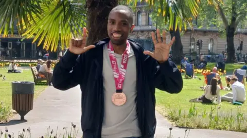 A man wearing a black hoodie, a beige sports top and a medal with a pink ribbon stands in front of a palm tree, in a park. He holds up seven fingers and smiles.