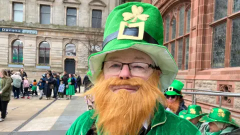 A man is dressed in costume as St Patrick. He is wearing a tall green hat with a shamrock on its front, a fake ginger beard , a green coat and also eye glasses. He is standing in front of a number of people in similar dress, while to his right a crowd of people can be seen