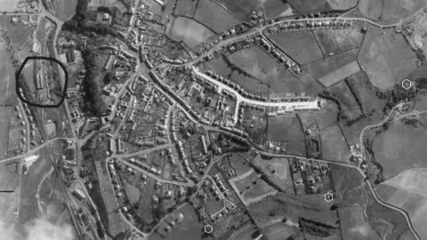 Supplied A black and white aerial image of Wooler taken in 1948 with a patchwork of houses and streets. On the left hand side two cylindrical buildings are circled. 