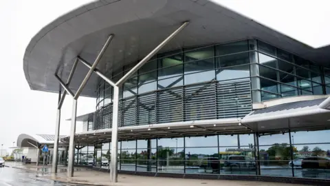 BBC Guernsey airport is a two-storey building made largely of glass and steel with a curved canopy roof is supported by three forked steel pillars. The windows are reflecting passing cars. It is raining.