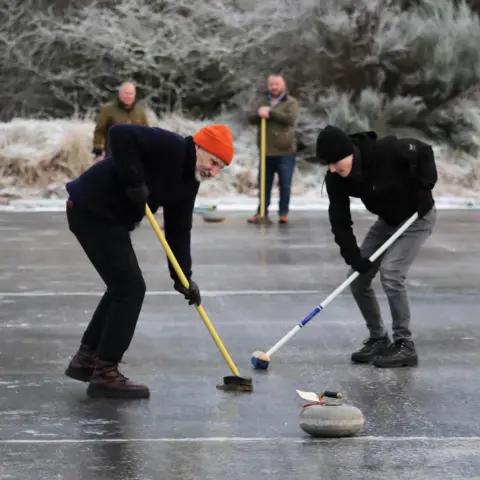 Peter Jolly/Northpix Two curler use brushes to brush the ice to help a curling stone travel down the rink. The men are wearing hats and gloves. Two other curlers watch from the sidelines.