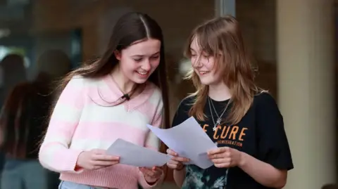 Two girls with brown hair holding their GCSEs smiling. One is wearing a pink and white jumper and one a black tshirt with orange writing on it. 