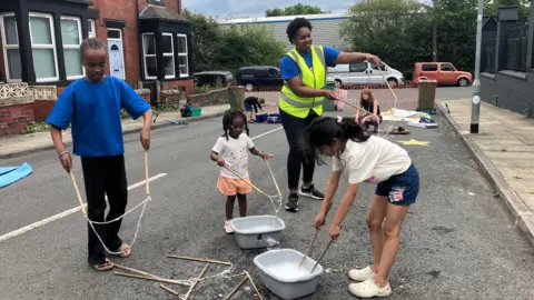 BBC Three girls are making large bubbles using a piece of string between two sticks. They are being watched by one female adult in a high vis vest who is doing the same thing. They are playing in the middle of a residential street which has been blocked off to traffic.