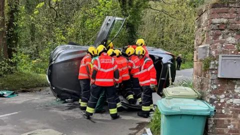Jersey Fire Service About eight firefighters work on a car which has flipped onto its side following a crash in St Peter's Valley, Jersey. The car is grey in colour. The garden wall of a property is next to the scene with two green wheelie bins propped up against the wall. Trees are on the opposite side.