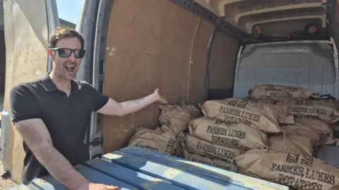 Luke, a man, with short brown hair. He is wearing a black T-shirt and black sunglasses and is gesturing towards brown sacks of potatoes in the back of a van. The bags all say "FARMER LUKES POTATOES".