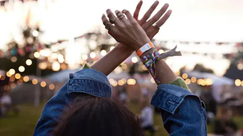 A woman with brown hair is wearing a denim jacket. She is at a festival with fairy lights and tents blurred in front of her. Her arms are in the air as if she is dancing. She has wristbands on her wrist and rings on her fingers