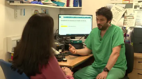 GP in green scrubs sitting at a desk talking to BBC reporter Philippa Goymer