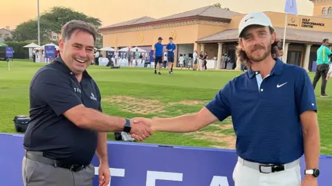 Norman Marshall Fleetwood and Marshall shaking hands in front of a sign on a golf green.